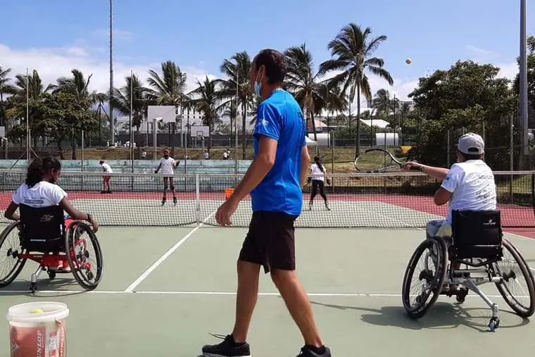 Un coach accompagne deux joueuses en fauteuil pendant qu'un groupe d'enfants s'entraîne sur le court voisin.